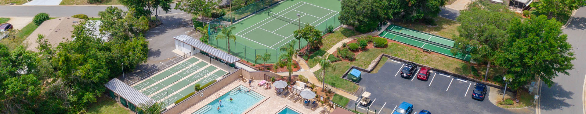Photo of tennis court, pools, parking lot, shuffle board courts, and trees taken from a drone in the sky looking down